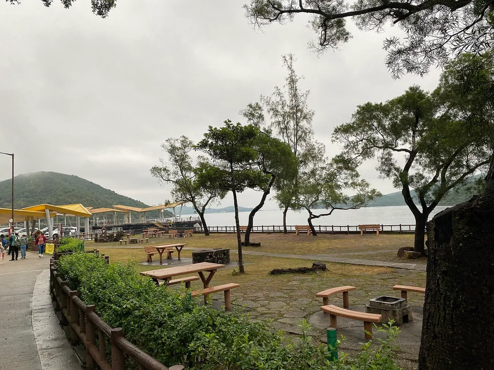 Picnic tables and barbecue pits under trees, with a small building behind.