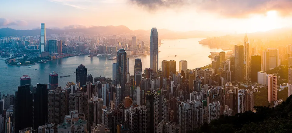 Panorama of Hong Kong Island skyscrapers and Victoria Harbour at sunrise, with Kowloon and hills beyond.