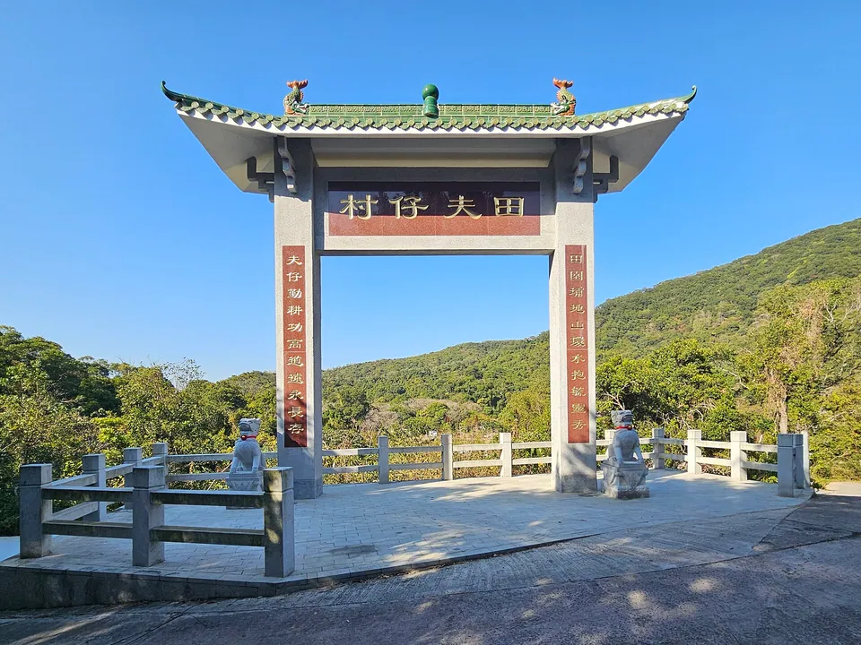 A white stone gateway with a green tiled roof and the name 田夫仔村, with wooded hills behind.
