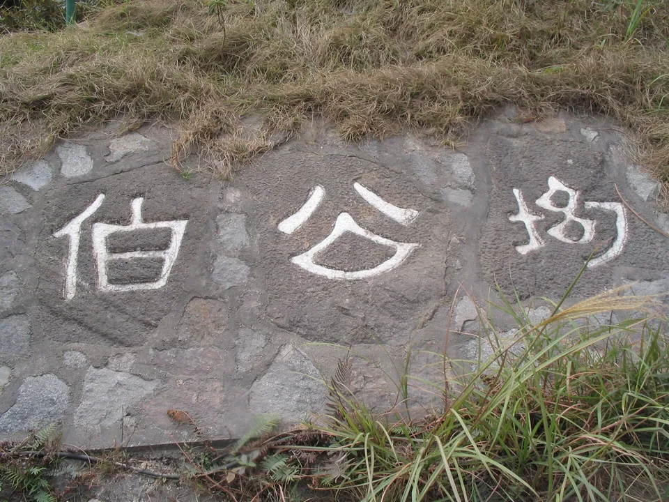 Large white Chinese characters 伯公坳 carved into a stone wall with grass above.