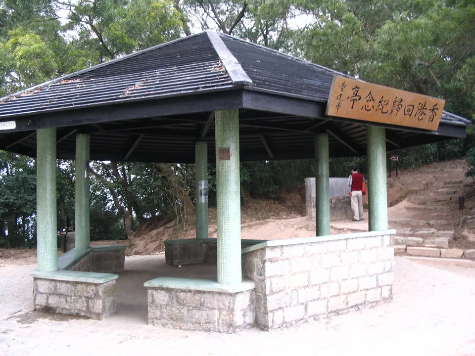 A square roofed pavilion with a wooden name board, stone seats and a path beside it.