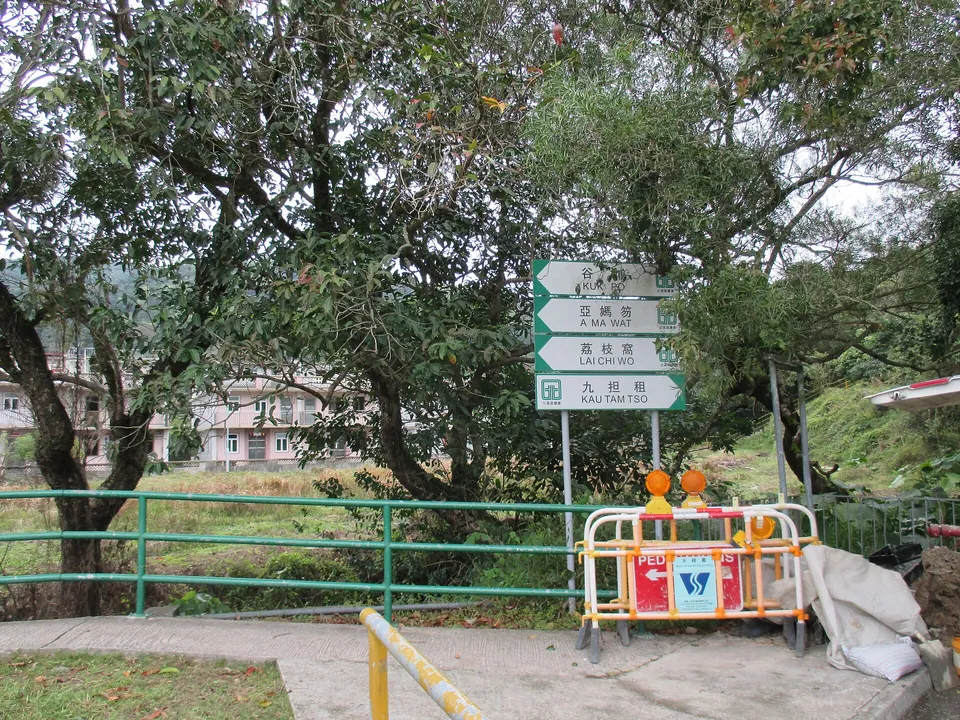 Green signposts to Kuk Po, A Ma Wat, Lai Chi Wo and Kau Tam Tso beside a railing, with village houses behind trees.