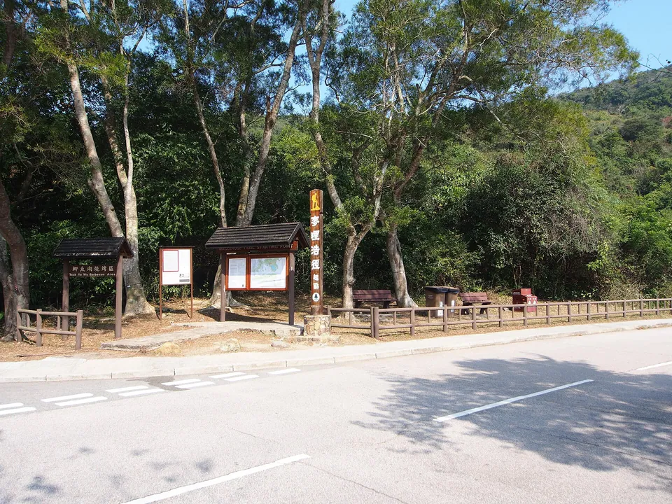 Roofed information boards and a tall trail marker post beside a road under trees.