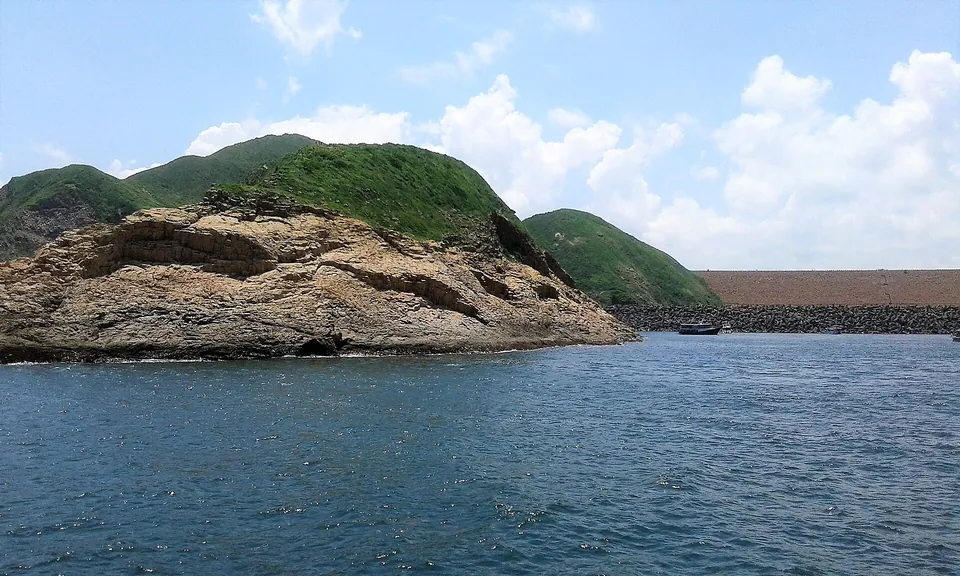 A rocky headland covered in green, with a long dam wall across the water behind it.